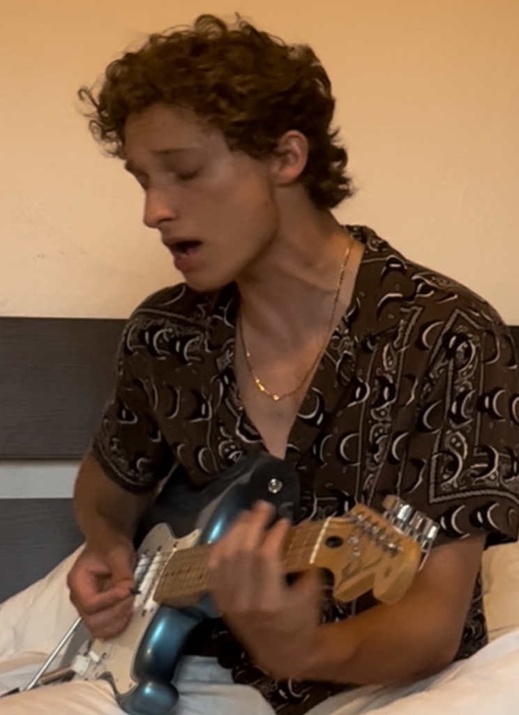 An image of the Author of the website, Sava from SavaStrums playing his electric guitar. He is a white man with dirty blond/brown hair. He is sat down in a bedroom with a loose brown patterned shirt and his bright blue guitar. His mouth is open as he sings and plays at the same time.