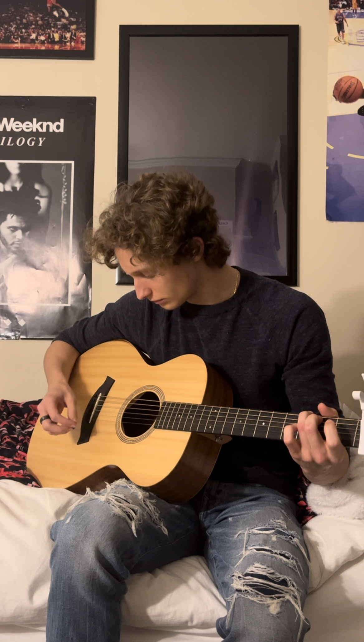 The author of SavaStrums (Sava), sitting in his bedroom playing the guitar. White man with dirty blond/brown hair looking down at his acoustic guitar strumming a chord. The guitar is a light-coloured wood, resting on his thigh and chest. The bedroom has many posters behind him, including one the Weeknd's trilogy album cover.