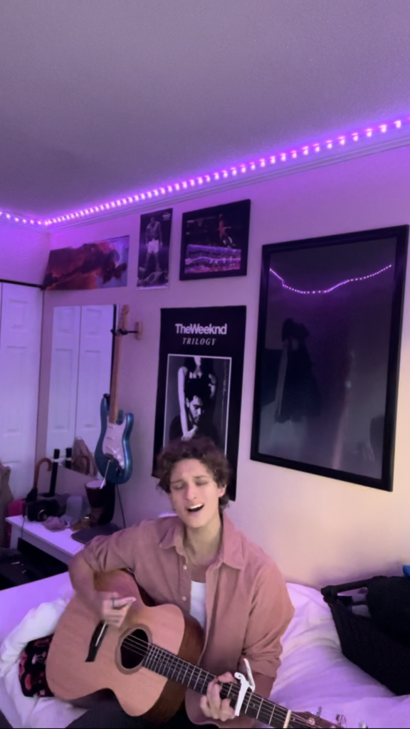 Image of the author of SavaStrums singing and playing the guitar in his bedroom sitting on his bed. His bedroom is scattered with posters of music albums and athletes. Additionally, the room is coloured a light purple via LED lights that are wrapped around the rooms ceiling corners. He is wearing a pink jacket and holding his bright, electric, blue guitar.