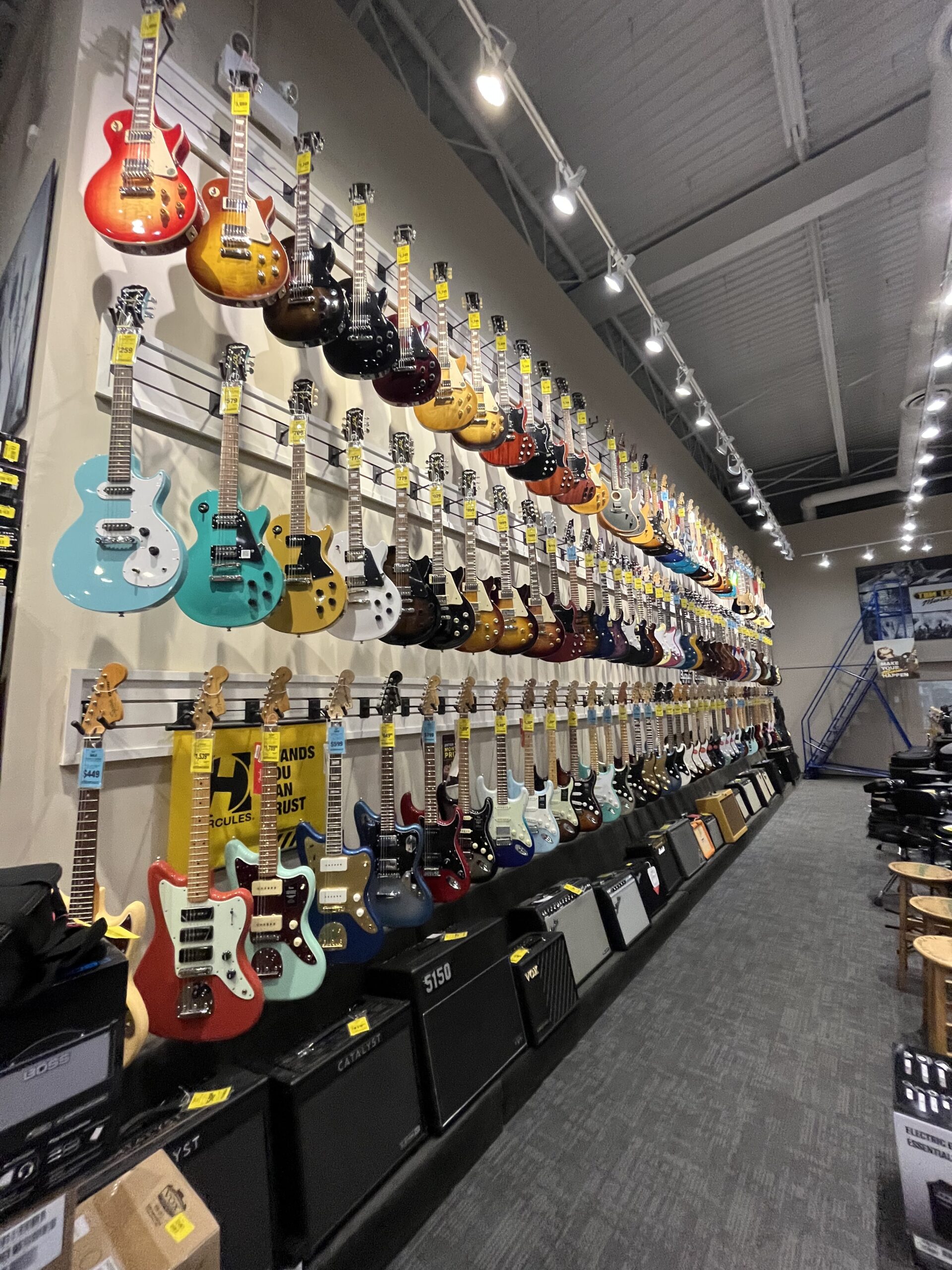 A wall of dozens upon dozens of electric guitars at a local guitar store. There are three rows of guitars that go up to the high ceiling, each different in colour. The picture is a splash of vibrancy displaying the beauty of a sguitar store and its electric guitars